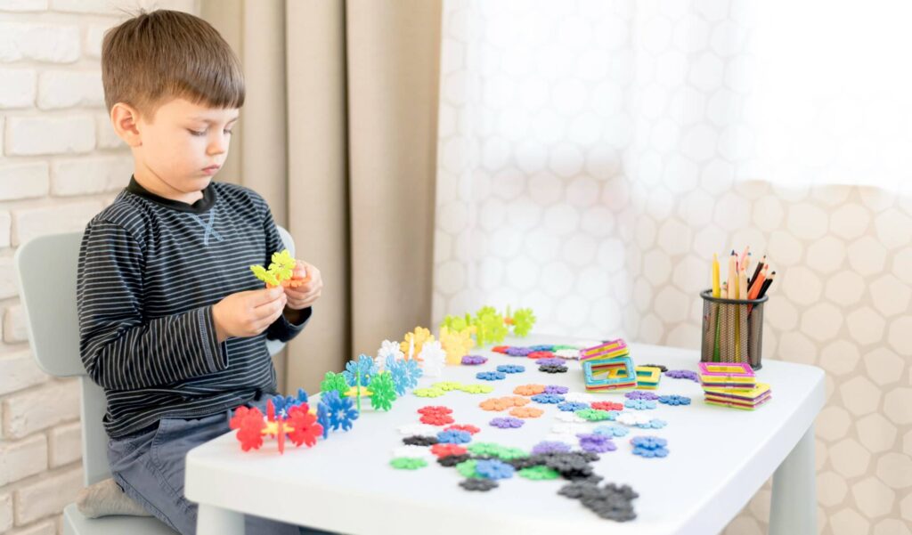 A young boy with short brown hair plays with colorful interlocking toys at a white table in a bright room.