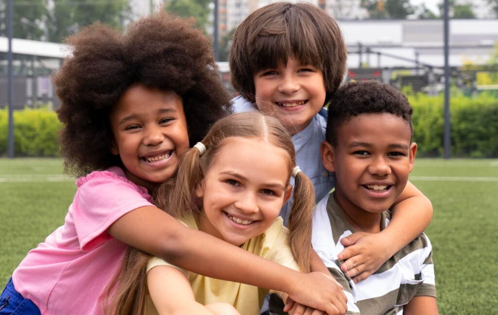 Four smiling children embrace on a green field, diverse in hair and skin tones, looking happy.