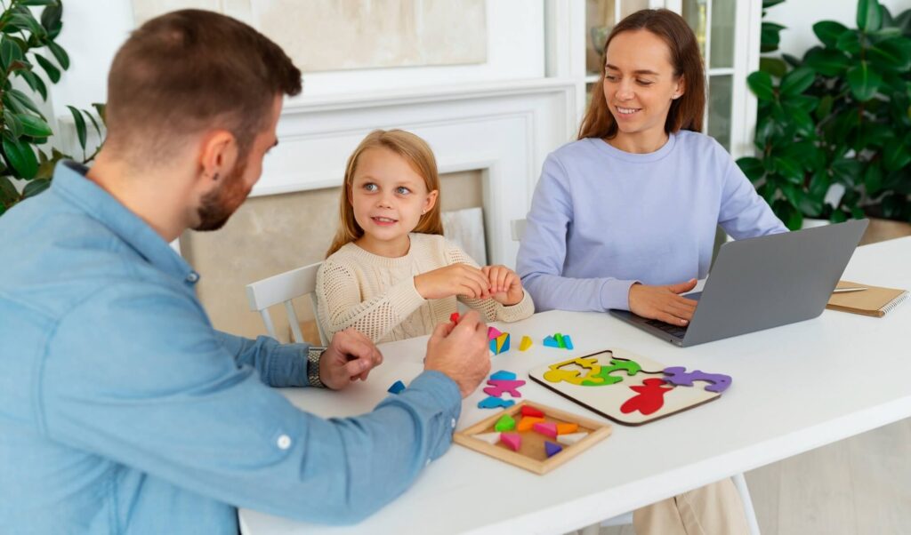 A family of three, including a young girl, works together on puzzles and a laptop at a bright white table.