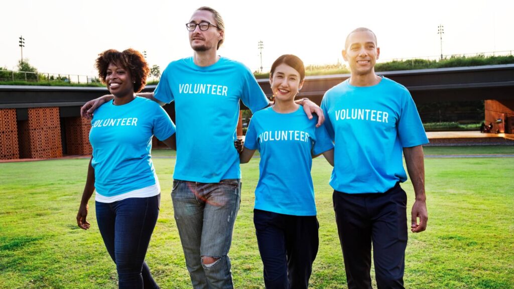 Four diverse volunteers in blue shirts walk arm-in-arm on a grassy field, smiling and looking directly at the viewer.